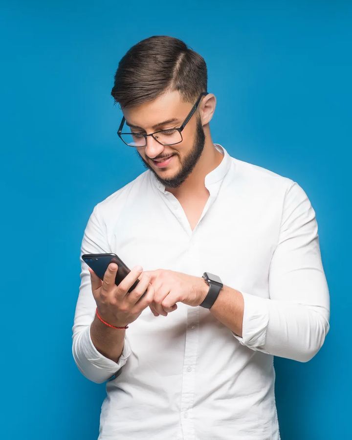Homem de camisa branca sorrindo enquanto usa o celular, sobre fundo azul de estúdio